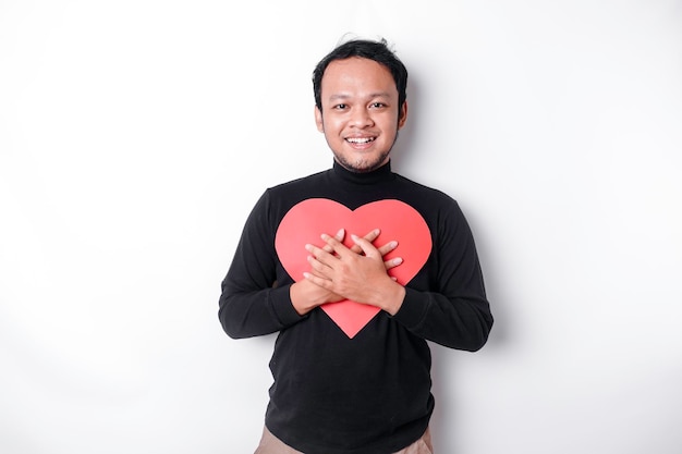 Un portrait d'un homme asiatique heureux portant une chemise noire tenant un papier en forme de coeur rouge isolé par fond blanc