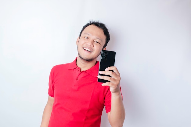 Un portrait d'un homme asiatique heureux sourit et tient son smartphone portant un t-shirt rouge isolé par un fond blanc