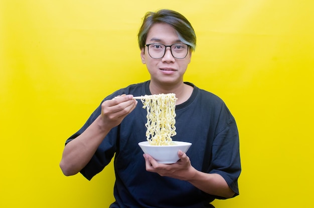 Portrait d'un homme asiatique heureux en t-shirt noir mange des nouilles instantanées à l'aide de baguettes et d'un bol isolés
