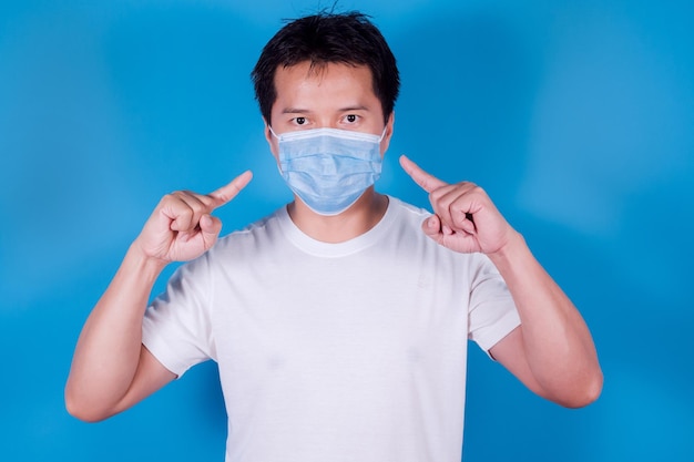 Portrait d'un homme asiatique pointant vers un masque médical pour protéger Covid-19 (Coronavirus) porte un t-shirt blanc sur fond bleu