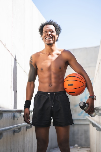 Portrait d'un homme athlète afro tenant un ballon de basket et se détendre après l'entraînement à l'extérieur. Sport et mode de vie sain.