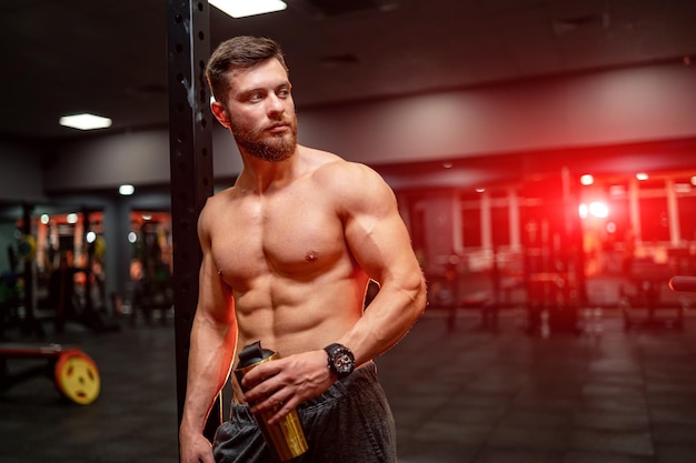 Portrait d'un homme athlétique bien bâti avec six pack abs. La moitié se tourna vers la caméra. Arrière-plan de la salle de sport. Guy tenant la bouteille. Fermer.