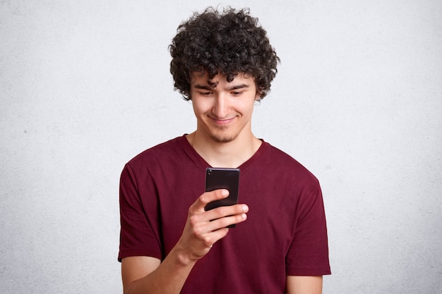 Portrait d'homme attrayant heureux aux cheveux bouclés et petite barbe tenant le smartphone et regardant son scren avec un sourire intrigué