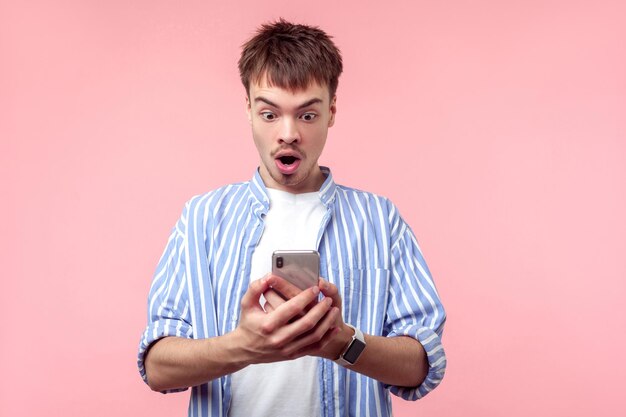 Portrait d'un homme aux cheveux bruns étonné avec une petite barbe et une moustache en chemise rayée décontractée lisant un message dans un téléphone portable étonné de nouvelles choquantes prise de vue en studio à l'intérieur isolée sur fond rose