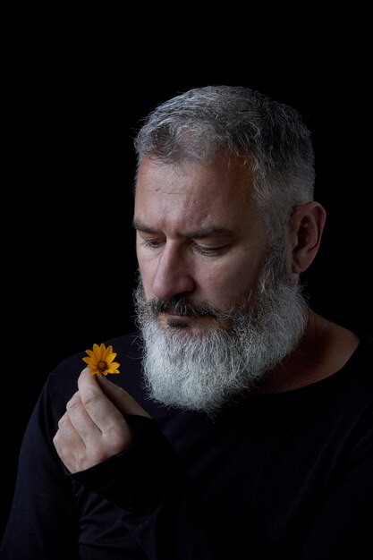 Portrait d'un homme aux cheveux gris brutal avec une barbe reniflant des fleurs jaunes sur fond noir, mise au point sélective