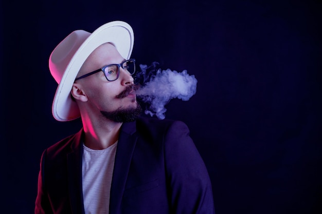 Portrait d'homme barbe en chapeau à larges bords blanc qui souffle de la fumée sur des filtres de couleur de fond sombre