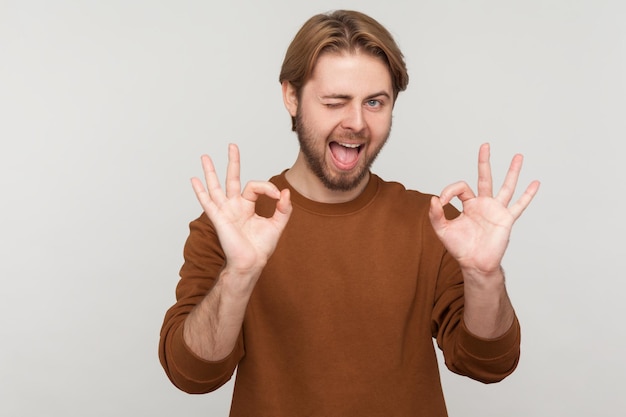 Portrait d'un homme à la barbe portant un sweat-shirt montrant un geste correct avec les doigts approuvant le travail satisfait de la qualité à la recherche d'un clin d'œil à la caméra Prise de vue en studio intérieure isolée sur fond gris