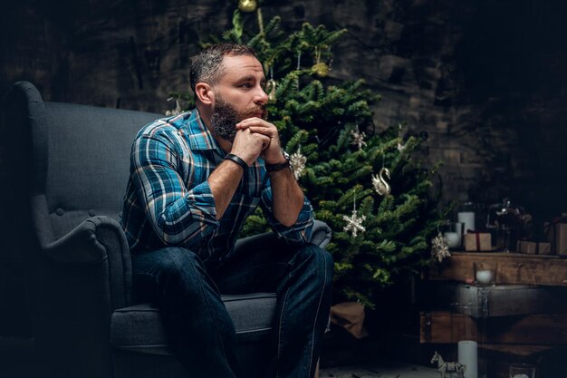 Portrait d'un homme barbu d'âge moyen vêtu d'une chemise en flanelle à carreaux et d'un jean est assis sur une chaise sur fond décoré de Noël.