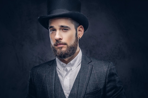 Portrait d'un homme barbu élégant dans un costume élégant et un chapeau cylindrique, regardant une caméra sur fond sombre.