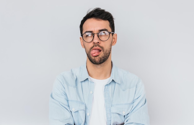 Portrait d'un homme barbu élégant porte des lunettes rondes à la mode et une chemise bleue a une expression dégoûtée avec la langue Expressions faciales humaines émotions concept de personnes et de sentiments