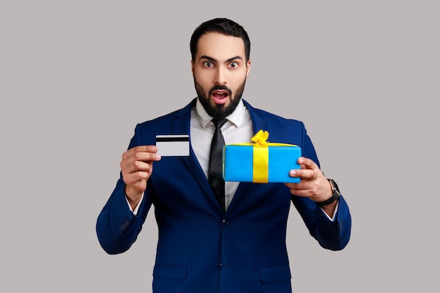 Portrait d'un homme barbu étonné et choqué montrant une carte de crédit et une boîte-cadeau, regardant avec étonnement, bancaire, portant un costume de style officiel. Tourné en studio intérieur isolé sur fond gris.