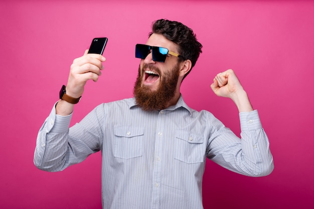 Portrait d'homme barbu étonné en tenue décontractée smartphone et célébrant la victoire