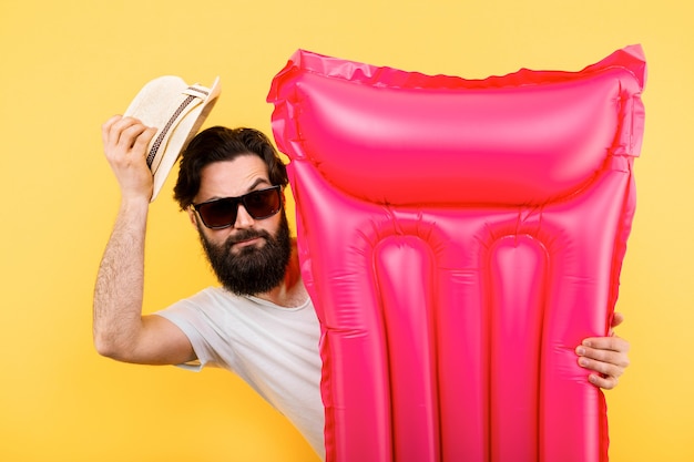 Portrait d'un homme barbu en lunettes de soleil et chapeau avec matelas de natation gonflable rose