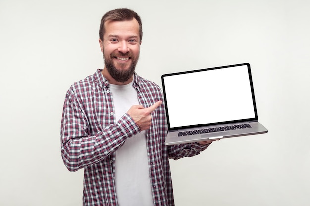 Portrait d'un homme barbu positif en chemise à carreaux décontractée tenant un ordinateur portable et pointant vers un écran vide souriant satisfait de l'application informatique studio d'intérieur tourné isolé sur fond blanc place pour l'annonce