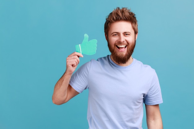 Portrait d'un homme barbu positif extrêmement heureux tenant et montrant un signe de forme de papier comme ou pouces vers le haut, regardant la caméra avec un sourire à pleines dents. Studio intérieur tourné isolé sur fond bleu.