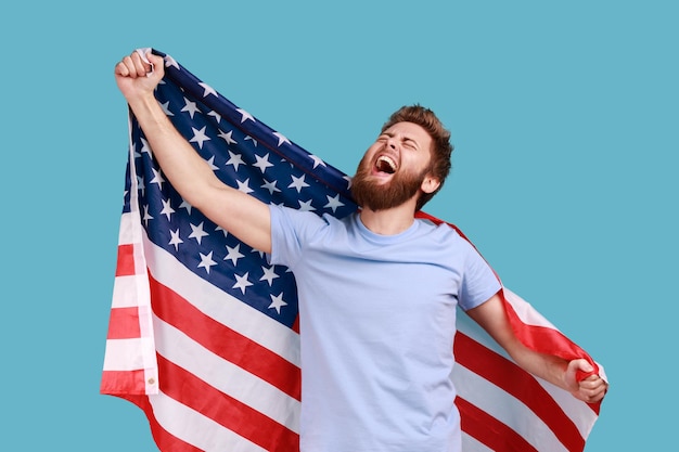 Portrait d'un homme barbu ravi et satisfait, tenant un énorme drapeau américain et se réjouissant tout en célébrant la fête nationale, levant les yeux et criant. Studio intérieur tourné isolé sur fond bleu.