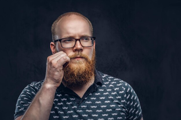 Portrait d'un homme barbu rousse pensif vêtu d'un t-shirt posant avec une main sur sa moustache. Isolé sur un fond texturé sombre.