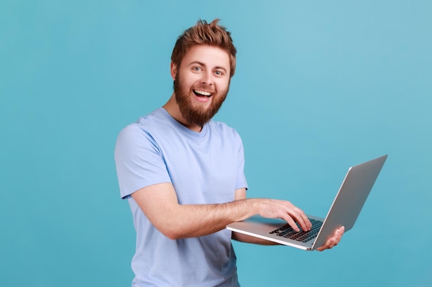 Portrait d'un homme barbu souriant tenant un ordinateur portable à la main et tapant un blogueur faisant des messages sur les réseaux sociaux discutant avec des adeptes regardant la caméra Prise de vue en studio intérieur isolée sur fond bleu