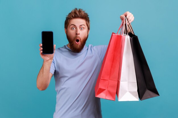 Portrait d'un homme barbu surpris étonné debout avec la bouche ouverte montrant des sacs à provisions et un téléphone intelligent avec écran vide pour la publicité. Studio intérieur tourné isolé sur fond bleu.