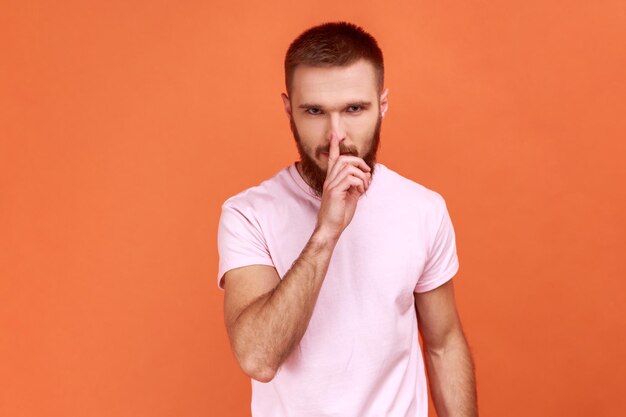Portrait d'un homme barbu tenant le doigt sur les lèvres faisant un geste de silence silencieux, demandant de garder le secret, ne parle pas, portant un T-shirt rose. Studio intérieur tourné isolé sur fond orange.