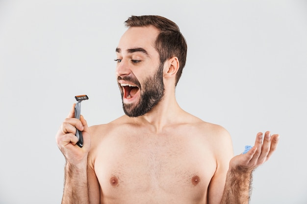 Portrait d'un homme barbu torse nu joyeux debout isolé sur blanc, tenant un rasoir