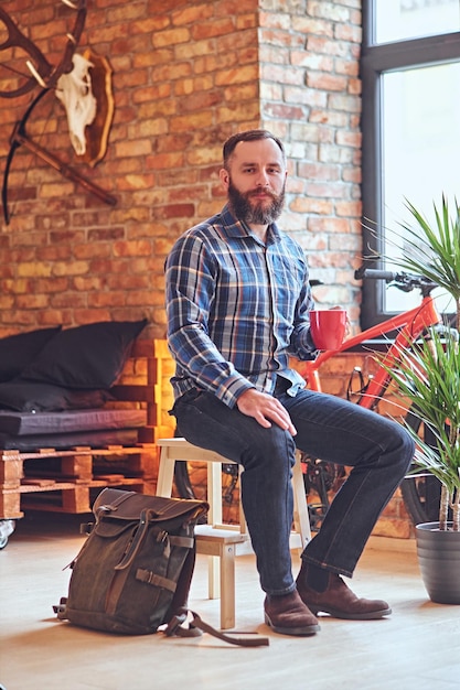 Portrait d'homme barbu vêtu d'une chemise polaire bleue, boit du café chaud près de la fenêtre dans la salle de repos avec intérieur loft.