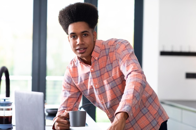 Portrait d'un homme biracial heureux, appuyé sur un comptoir, buvant du café, utilisant un ordinateur portable dans une cuisine ensoleillée