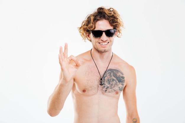 Portrait d'un homme bouclé souriant en short de plage montrant le geste correct isolé sur le mur blanc