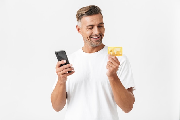 Portrait d'un homme brune des années 30 portant un t-shirt décontracté tenant un smartphone et une carte de crédit en plastique isolée sur blanc