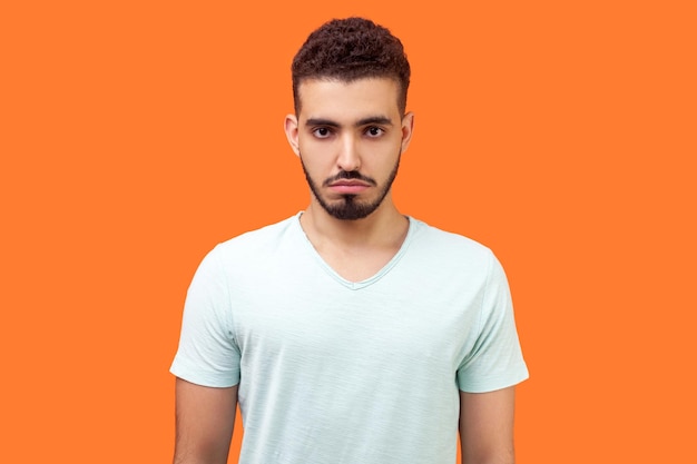 Portrait d'un homme brune bouleversé avec une barbe en t-shirt blanc décontracté regardant la caméra avec une expression de visage insatisfait et plein de ressentiment mauvaise humeur dépression prise de vue en studio à l'intérieur isolée sur fond orange