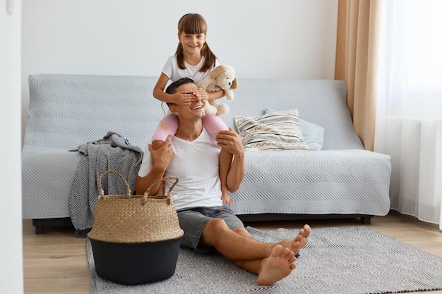 Portrait d'un homme brune vêtu d'un t-shirt blanc de style décontracté et d'un jean court assis sur le sol tenant une petite fille sur ses épaules fille couvrant les yeux des papas avec les paumes