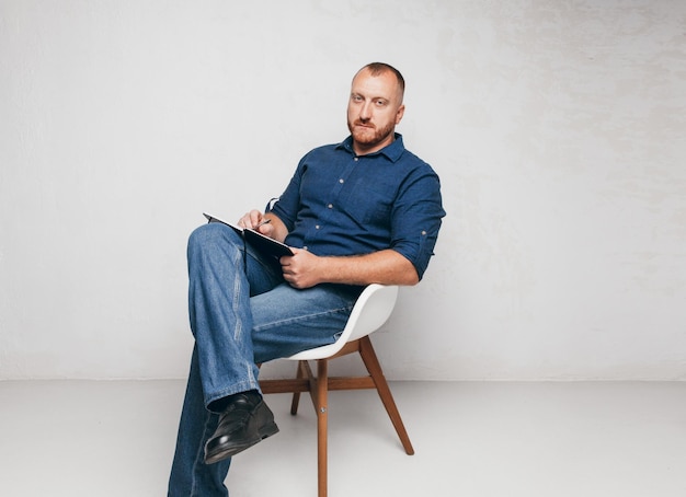 Portrait d'un homme brutal assis sur une chaise contre un mur blanc avec un ordinateur portable et un homme d'affaires bloc-notes