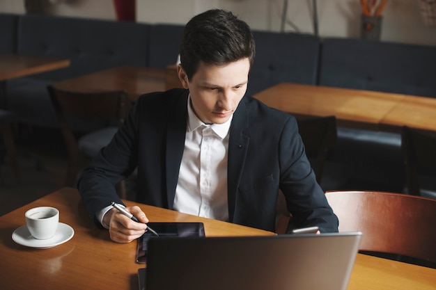 Portrait d'un homme caucasien élégant habillé en costume en regardant son ordinateur portable tout en fonctionnant sur une tablette assis à son bureau dans un café.