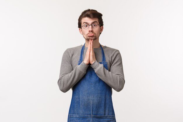 Portrait d'homme caucasien idiot mignon en tablier, employé de café demandant à un collègue de le couvrir au travail, besoin de rentrer tôt, dites s'il vous plaît avec les mains dans la prière,