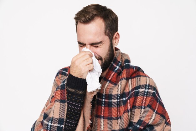 Portrait d'un homme caucasien malade enveloppé dans une couverture éternuant et tenant une serviette en papier à cause de la grippe isolée sur un mur blanc