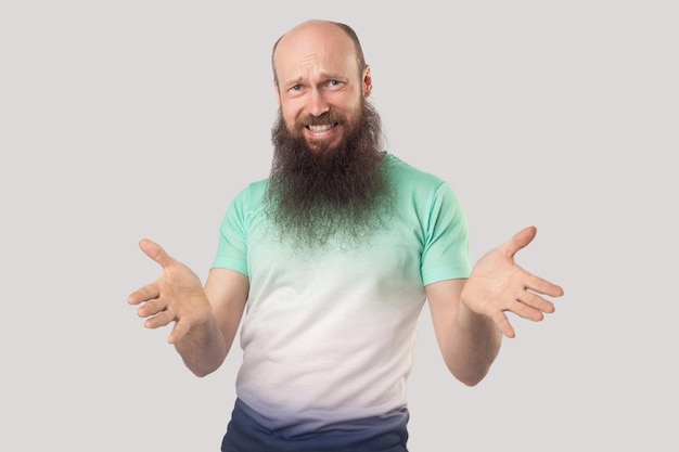 Portrait d'un homme chauve d'âge moyen en colère avec une longue barbe en t-shirt vert clair debout, regardant la caméra avec les bras levés et demandant. studio d'intérieur tourné isolé sur fond gris