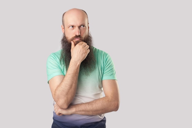 Portrait d'un homme chauve d'âge moyen confus avec une longue barbe en t-shirt vert clair debout avec la main sur le menton et regardant ailleurs et pensant quoi faire. tourné en studio intérieur, isolé sur fond gris.