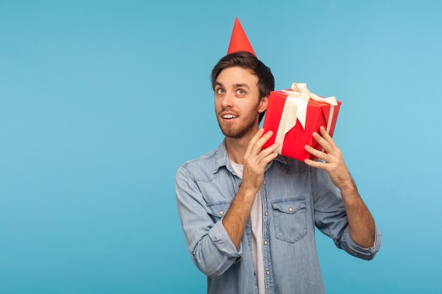 Portrait d'un homme curieux avec un chapeau de fête drôle tenant une boîte-cadeau près de l'oreille et écoutant, devinant le cadeau, en prévision d'une surprise d'anniversaire intéressante. studio d'intérieur tourné isolé sur fond bleu