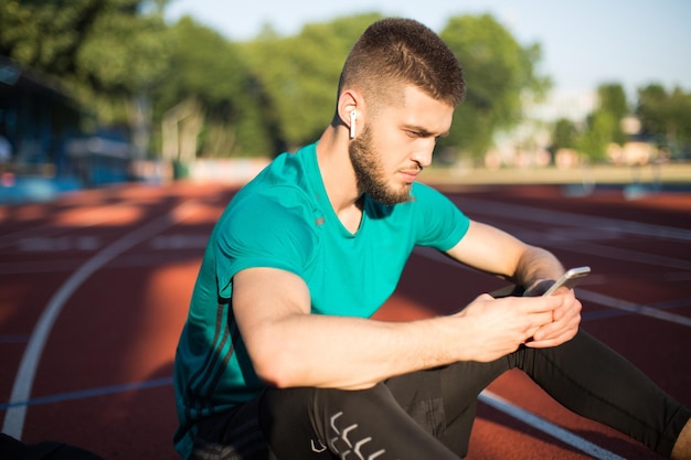 Portrait d'un homme dans des écouteurs sans fil utilisant judicieusement un téléphone portable tout en passant du temps sur la piste de course du stade