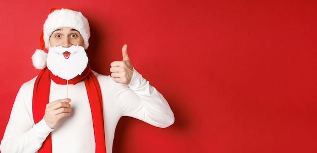 Portrait d'un homme drôle en bonnet de noel tenant un masque de barbe montrant le pouce vers le haut profitant de la fête du nouvel an debout sur fond rouge