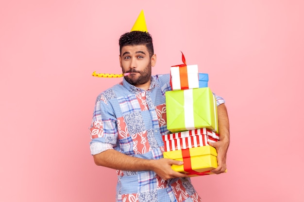 Portrait d'un homme drôle et heureux avec une barbe portant une chemise de style décontracté et un cône tenant une pile de cadeaux et soufflant une corne de fête célébrant l'anniversaire Prise de vue en studio intérieure isolée sur fond rose