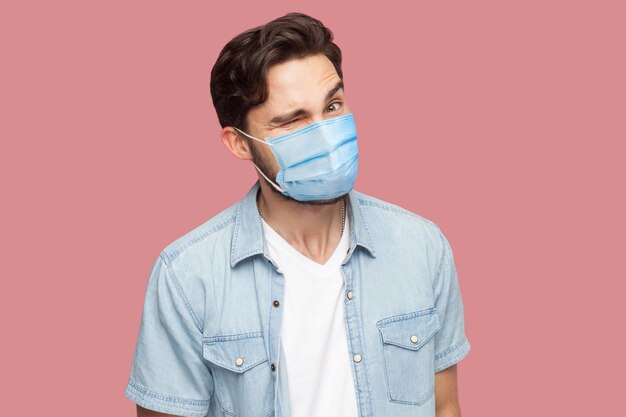 Portrait d'un homme drôle avec un masque médical chirurgical en chemise bleue de style décontracté debout, un clin d'œil et regardant la caméra avec une grimace et un sourire. tourné en studio intérieur, isolé sur fond rose.