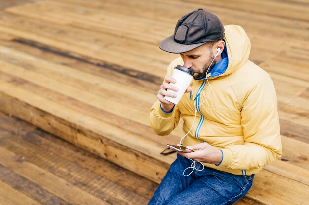 Portrait d'un homme élégant avec une barbe portant une veste jaune et un jean, buvant un délicieux café et écoutant de la musique avec des écouteurs tenant un téléphone portable téléchargeant des photos et des vidéos en ligne