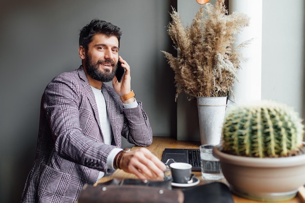 Portrait d'un homme élégant barbu utilisant un ordinateur portable au café tout en étant assis à la table en bois, en buvant du café et en parlant par téléphone