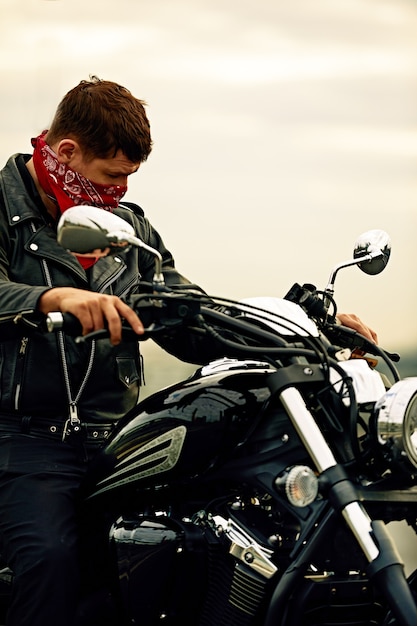 Portrait d'un homme élégant vêtu d'une veste en cuir noir et d'un bandana assis sur sa moto rétro sur mesure, détournant les yeux, bâtiments de la grande ville en arrière-plan.