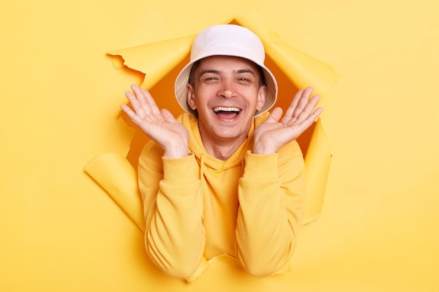 Portrait d'un homme étonné excité heureux portant un sweat à capuche décontracté et un panama regardant à travers un trou déchiré dans du papier jaune regardant la caméra avec un sourire heureux bras levés exprimant des émotions positives