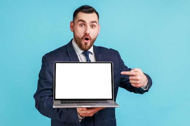 Portrait d'un homme excité surpris avec une barbe en costume pointant du doigt un écran d'ordinateur portable vide regardant avec de grands yeux choqués par la publicité. Studio intérieur tourné isolé sur fond bleu.