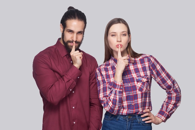 Portrait d'un homme et d'une femme barbus sérieux dans un style décontracté debout et regardant la caméra avec les doigts sur les lèvres et montrant un signe silencieux. tourné en studio intérieur, isolé sur fond gris.