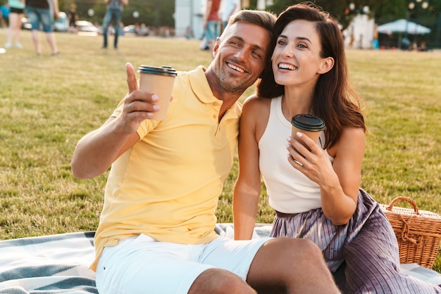 Portrait d'un homme et d'une femme excités d'âge moyen buvant du café à emporter et pointant du doigt tout en étant assis sur l'herbe dans le parc