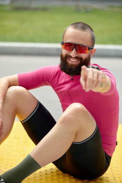 Portrait d'un homme de fitness souriant se reposant assis sur un sol sur le terrain d'athlétisme à l'extérieur, pointant son doigt vers la caméra